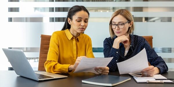 Two,Diverse,Businesswomen,Reviewing,Documents,And,Discussing,Work,,Collaborating,On